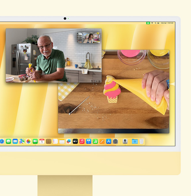 Gele iMac. Demonstratie van de feature Bureauweergave. Op een scherm is een bakker te zien die met iemand aan het videobellen is. In het ene venster zijn de bakker en het gezicht van de man te zien. En in het andere zijn de handen van de bakker zichtbaar tijdens het glazuren van een koekje.