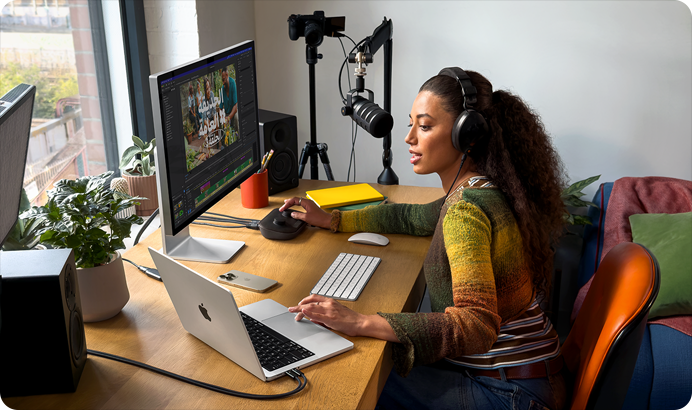 امرأة في منزلها تعمل خلف مكتبها على MacBook Pro متصل بشاشتين خارجيتين وترتدي سماعات رأس وتتحدث في الميكروفون