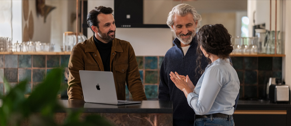 Dois homens e uma mulher conversam enquanto um MacBook está em cima de uma mesa.