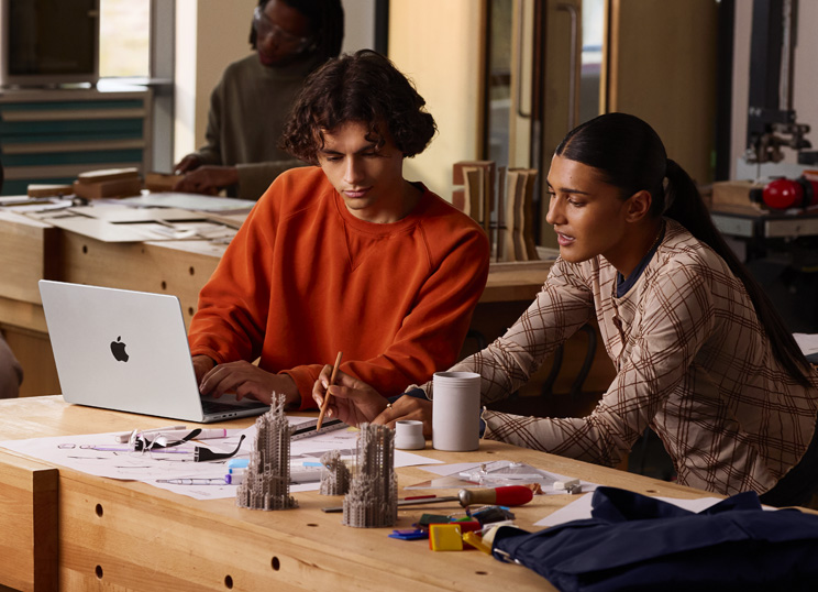 周りに画材が置かれたテーブルで、2人の人物が共同でプロジェクトに取り組んでいる。1人はMacBookにタイプ入力し、もう1人は紙の上で鉛筆と定規を使って作業している