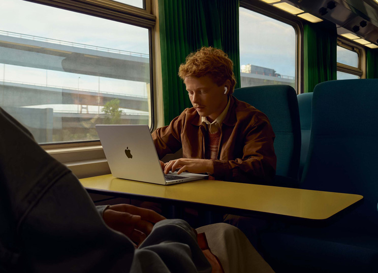 電車の窓際の席で、AirPodsを装着してMacBookを操作している人物