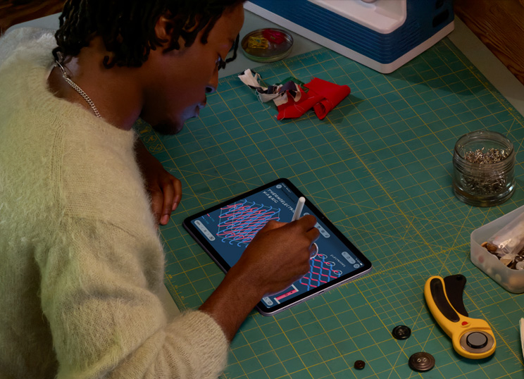 裁縫道具が置かれた緑色のカッティングマットの前に座り、iPadとApple Pencilを使ってイラストを描いている人物