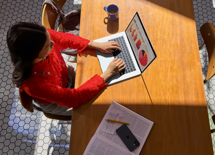 テーブルでMacBookを使っている人物。そばにマグカップ、スマートフォン、書類が置かれている