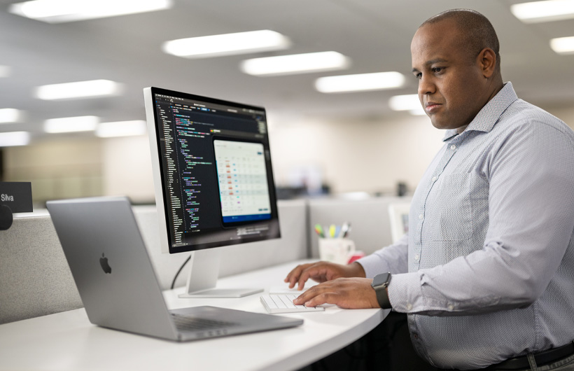 Een ontwikkelaar staat aan zijn bureau en werkt met een Mac en MacBook, op het Mac-scherm is code en app-informatie te zien